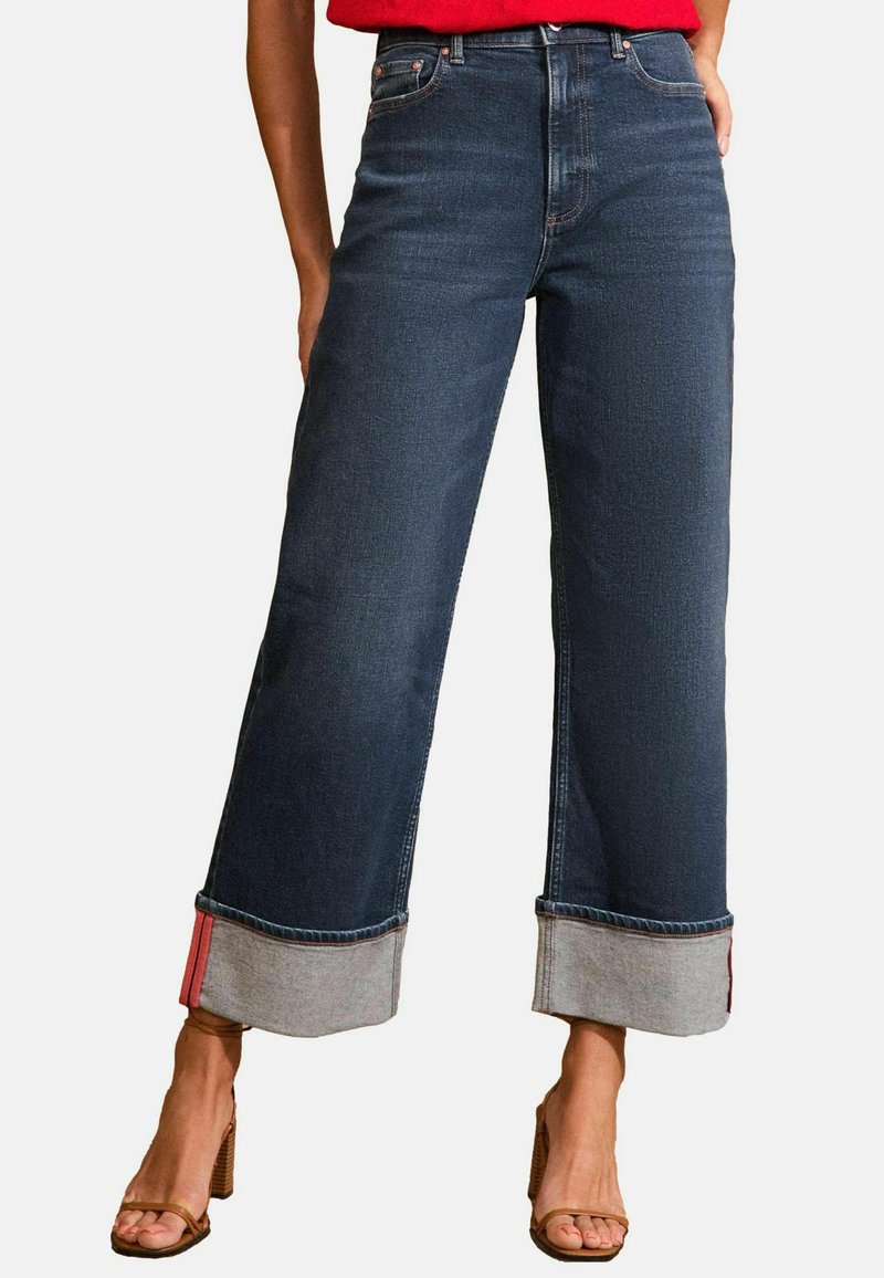 Farkut tummasta denimmateriaalista, jossa on korkea vyötärö ja leveät lahkeet. Mallissa on kontrastiväriset harmaat lahkeen suut ja punaiset korostusompeleet. Kankaan pinta on sileä ja klassinen viisi-taskumalli.