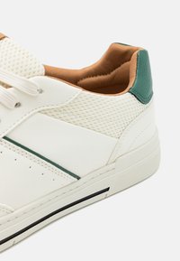 Sneaker bianca con pannelli in rete perforata, accenti verdi sul tallone e rivestimento interno beige. Dettaglio suola nera.