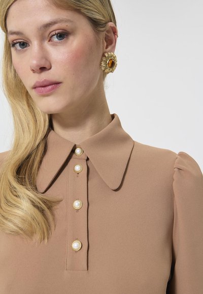 Beež blouse klassikalise kraega, millel on punnis varrukad ja kuldsed nupud, millele on lisatud pärleid. Kanda suurt lillemotivi kõrvarõngast.