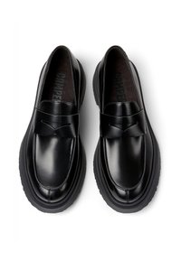 Mocasines de cuero negro con suelas de goma gruesas, puntas redondeadas y una banda decorativa en la parte delantera, vistos desde arriba sobre un fondo blanco.