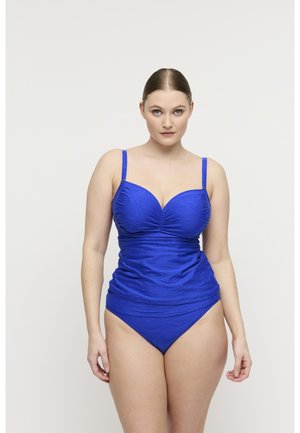 Vrouw met een lichte huid en blauwe ogen, gekleed in een blauw textuur eenstukbadpak, staand tegen een effen witte achtergrond.