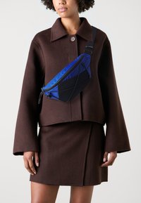 Blaue und schwarze Stoff-Schultertasche mit geschwungener Form, Rei�ßverschluss und verstellbarem Riemen, getragen über einem braunen oversize Mantel.