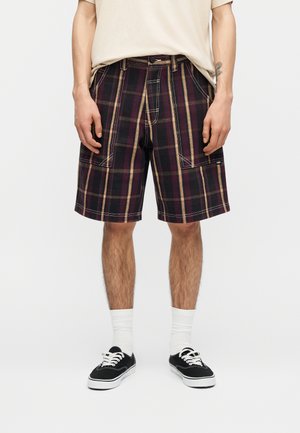 Uomo che indossa una maglietta beige, pantaloncini a quadri scuri fino al ginocchio con tasche frontali, calzini bianchi e scarpe da ginnastica nere con lacci.