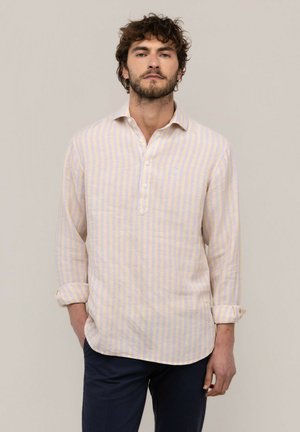 Hombre con cabello rizado castaño y barba, vistiendo una camisa de rayas blancas y amarillo claro de manga larga, con las manos en los bolsillos, de pie frente a un fondo liso.