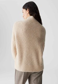 Beige gebreide trui met een gestructureerd, geribbeld patroon; heeft een ontspannen pasvorm en hoge kraag. Draag over grijs geruite broek.