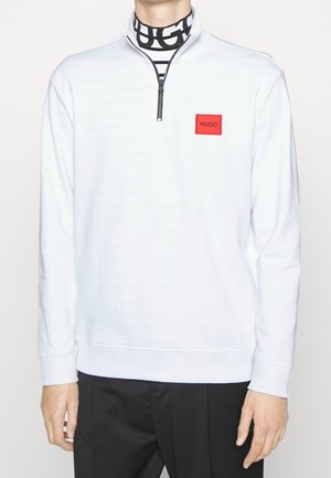 Mann trägt ein weißes Half-Zip-Sweatshirt mit rotem HUGO-Logo-Patch auf der Brust und einen schwarz-weiß gestreiften Rollkragenpullover.