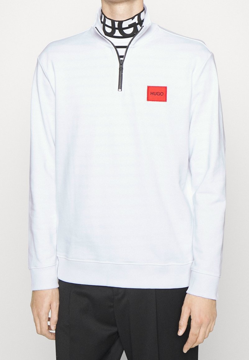 Homme portant un sweat-shirt blanc à demi-fermeture éclair avec un patch logo HUGO rouge sur la poitrine et un col roulé rayé noir et blanc.