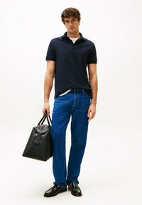 Marine polo shirt, blauwe jeans en zwarte leren schoenen. Houdt een gestructureerde zwarte tas vast. Eenvoudig ontwerp met een getailleerde pasvorm.
