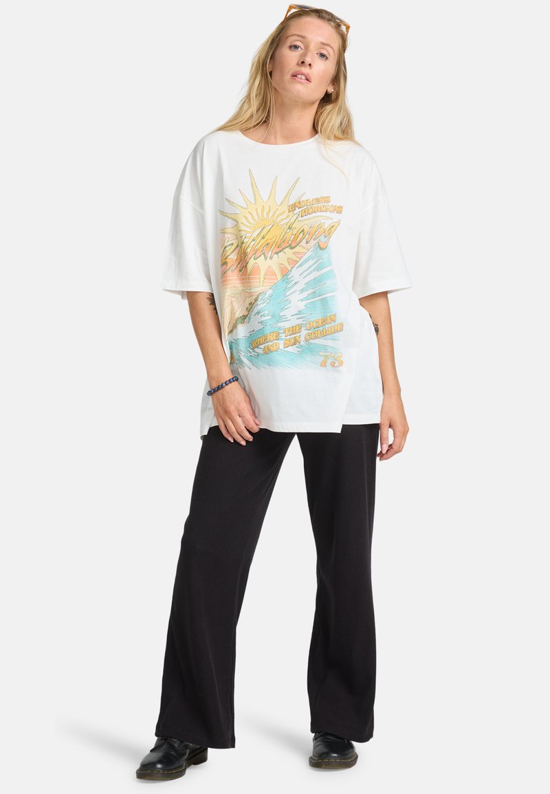 White oversized t-shirt with a colorful graphic of a sun and waves. Paired with black wide-leg pants and black shoes. Casual design.