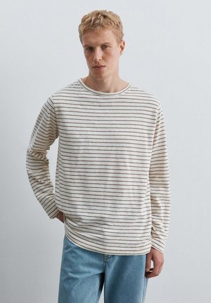 Lyshåret mand står op ad en ensfarvet væg iført hvid langærmet skjorte med tynde, vandrette brune striber og lyseblå jeans, den ene hånd i lommen.