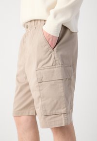 Calções cargo bege com um corte relaxado, cintura elástica, bolsos laterais e um bolso de remendo na coxa para armazenamento adicional.