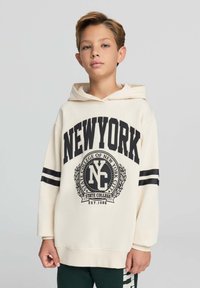 Felpa crema con la scritta "NEW YORK" in grassetto nero. Presenta un'emblema circolare sul petto, strisce nere sulle maniche e una vestibilità comoda.
