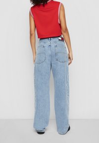 Jeans de pierna ancha de color azul claro con una cintura alta, que presentan un diseño de logo blanco en el lateral y una textura clásica de denim. Se ven zapatos negros.
