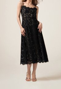 Robe midi en dentelle noire avec des bords festonnés, des bretelles, des motifs floraux et une silhouette évasée, associée à des talons roses en or à lanières.