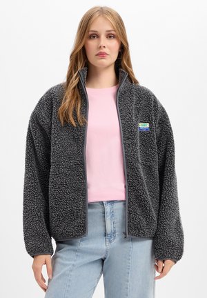 Frau mit langen blonden Haaren trägt eine dunkelgraue Fleecejacke über einem rosa Shirt und hellblaue Jeans, steht vor einem schlichten Hintergrund.