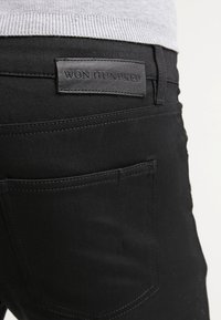 Jean en denim noir avec une étiquette en cuir portant la mention "WON HUNDRED", avec des poches arrière et une texture lisse, partiellement visible.