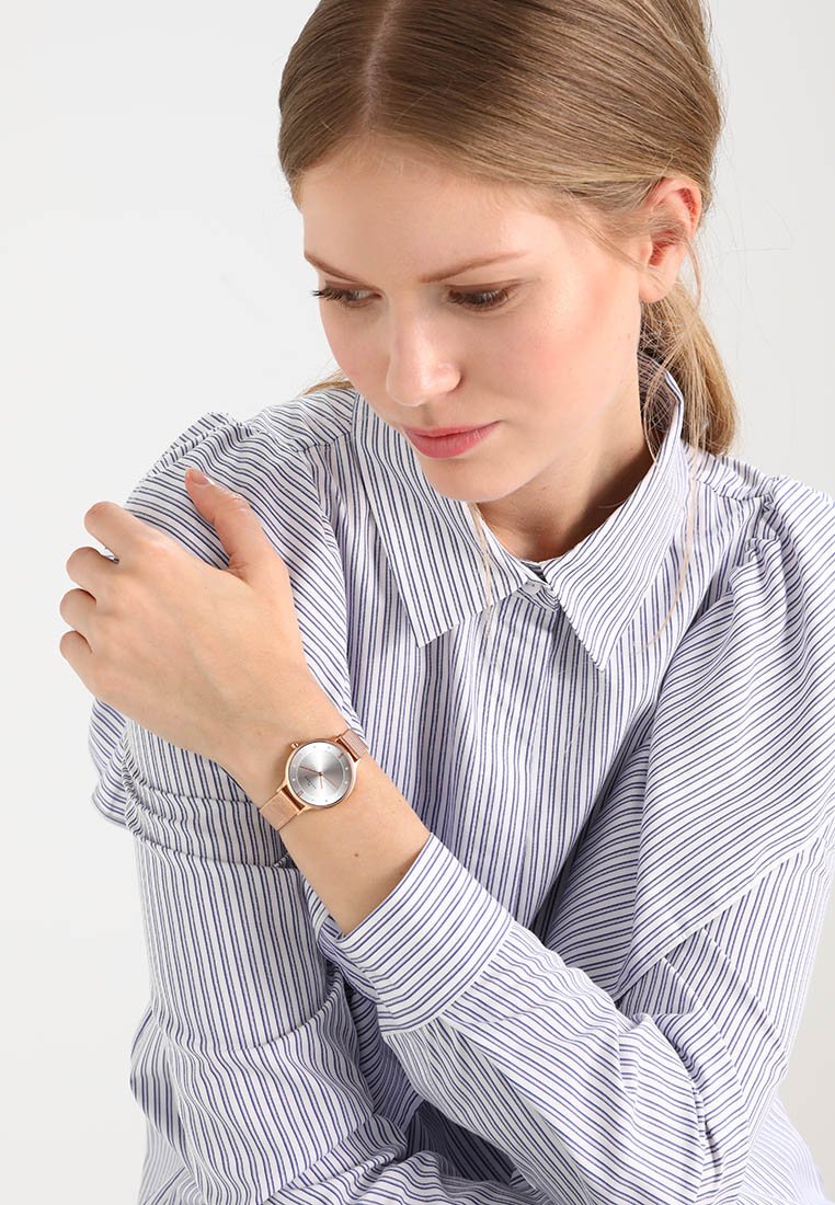 Un reloj plateado con una esfera redonda y acentos en oro rosa, combinado con una camisa de botones a rayas azul y blanca con cuello y mangas texturizadas.