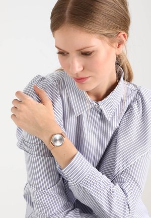 Frau mit hellbraunen Haaren, die ein gestreiftes Hemd mit Knöpfen trägt, blickt auf ihr Handgelenk mit einer minimalistischen silbernen Uhr mit einem rosegoldenen Armband.