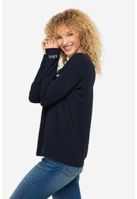 Marineblaue Langarm-T-Shirt mit dem Aufdruck "LOVE" in Weiß auf dem Ärmel; kombiniert mit blauen Jeans. Weicher Stoff, lockere Passform und minimales Design.