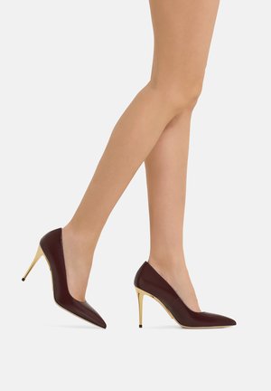 Gambe in collant nudo trasparente che indossano décolleté scuri bordeaux con punta affilata e tacchi a spillo dorati lucidi su sfondo bianco.