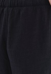 Pantalones de lino negros con cintura elástica. La tela tiene un acabado texturizado y un corte relajado, mostrando sutiles detalles de tejido.