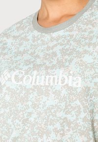 Ljusblå t-shirt med ett strukturerat, fläckigt mönster i grått och krämfärgat. Ordets "Columbia" framhävs tydligt i vitt.