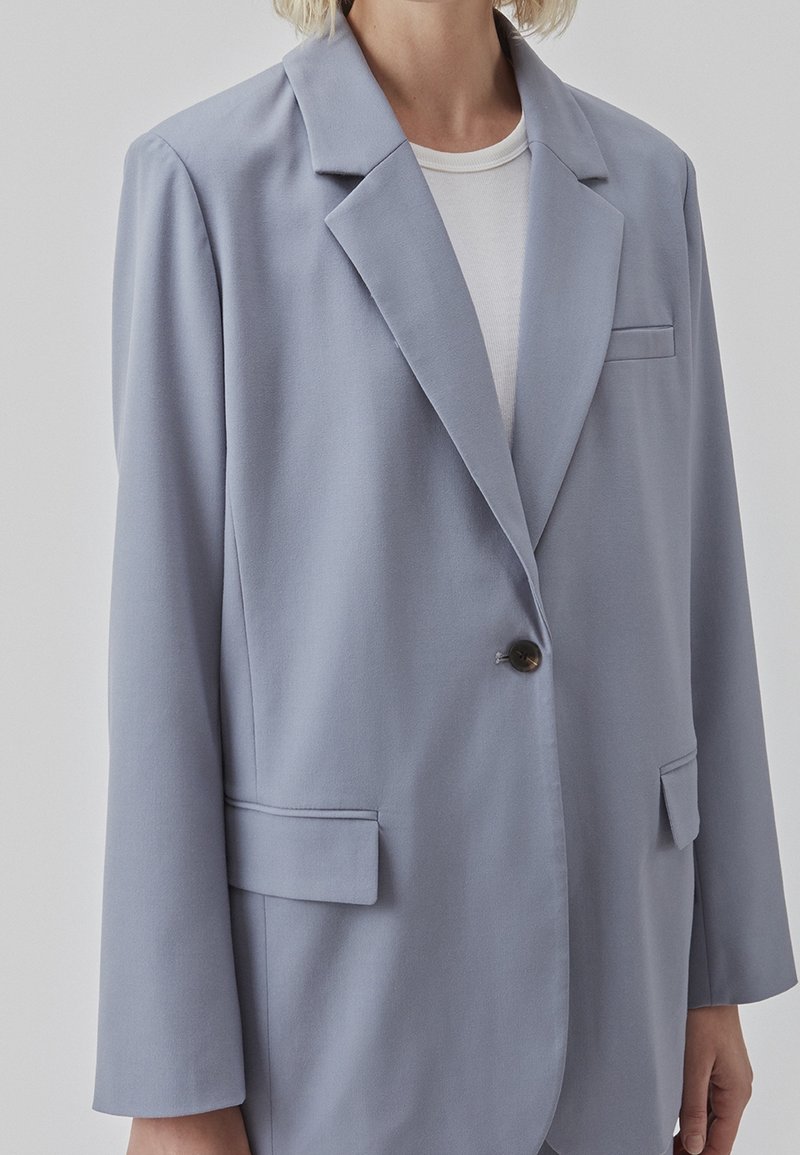 Blazer gris clair, design à un bouton, revers structurés, deux poches frontales, fabriqué en tissu lisse avec une légère brillance.