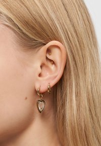 Blond öra med tre guldörhängen: en liten hoops, en medelstor hoops med en transparent hjärtanhänge, och två guldstuds på insidan av örat.