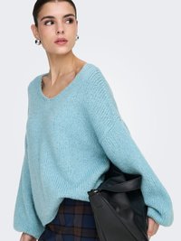 Vaaleansininen neulepaita leveällä pääntiellä ja pitkillä, pussimaisilla hihoilla. Yhdistetty mustaan nahkalaukkuun ja ruudulliseen hameeseen. Pehmeä rakenne.