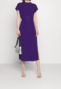Fialové maxi šaty s krátkým rukávem, uzlovým detailem na boku a bočním rozparkem. Nosené se stříbrnou kabelkou a prostornými sandály na podpatku.