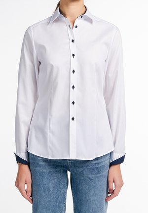 Femme portant une chemise blanche cintrée à boutons sombres et un jean bleu, debout devant un fond blanc uni.