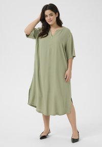 Vestido largo de manga corta en una suave y ligera tela verde. Presenta un cuello simple y aberturas laterales. Se lleva con unos zapatos planos negros de punta.