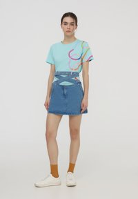 T-Shirt grafico turchese con linee astratte colorate, abbinato a una gonna di jeans blu stretta in vita con una cintura coordinata.