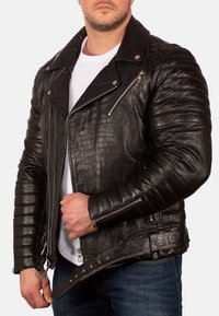 Zwarte leren motorjack met krokodillenstructuur, asymmetrische rits, schouderepauletten en gevoerde mouwen. Gestyld over een wit shirt.