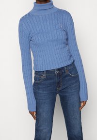 Suéter azul claro de punto trenzado con cuello alto y mangas largas, combinado con jeans azul oscuro. Detalles notables incluyen un pequeño logotipo en el pecho.