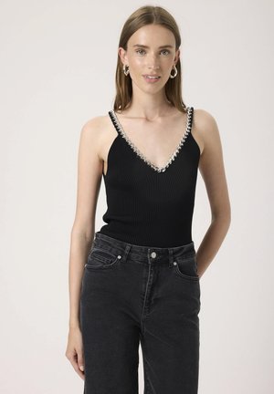 Jonge vrouw die een zwart geribd tanktop draagt met zilveren kettingdetail en een zwarte high-waisted jeans, staand tegen een effen achtergrond.