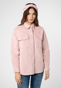 Lichtroze corduroy overhemdjasje met een gekraagde halslijn, twee voorzakken en knoopsluiting. Draagt een bijpassende haarband.