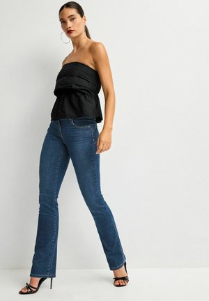 Top nero senza spalline con design plissettato e chiusura a bottone, abbinato a jeans svasati blu scuro e sandali neri con tacco alto.