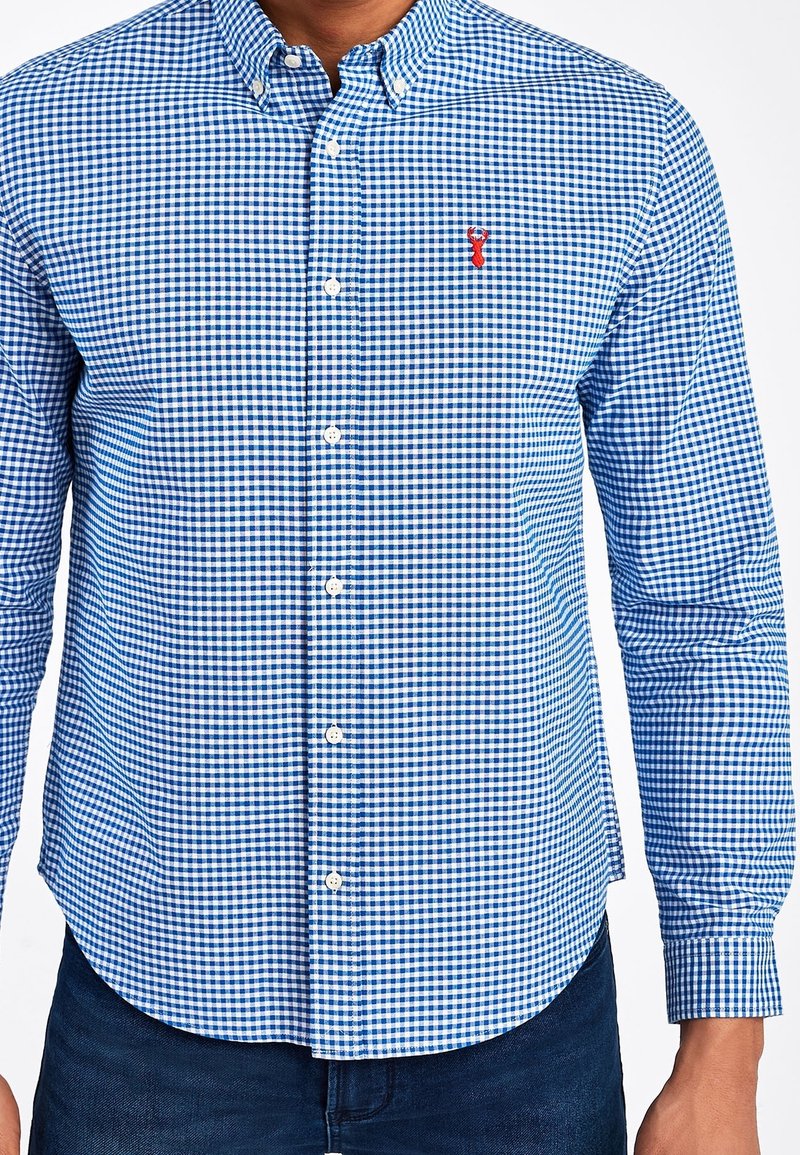 Camicia a quadri blu e bianca con maniche lunghe, colletto con bottoni e orlo arrotondato; presenta un piccolo logo rosso sul petto.