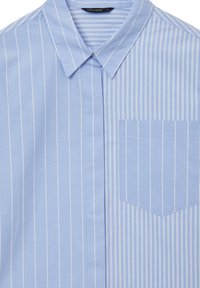 Camicia azzurro chiaro con righe verticali bianche, colletto a punta e taschino a sinistra. Tessuto morbido con una consistenza liscia.