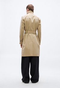 Béžový trenčkot so zapínaním v páse opaskom, zadným rozparkom a gombíkovými manžetami, v kombinácii s voľnými čiernymi nohavicami, pohľad zozadu.