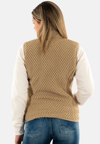 Chaleco texturizado color canela con un patrón acolchado, que presenta un cuello alto, diseño sin mangas y está unido a una camisa de manga larga de un color claro.