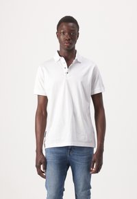 Witte poloshirt met een drieknopenlijst, korte mouwen en subtiel logo op de borst, gecombineerd met lichtblauwe jeans.