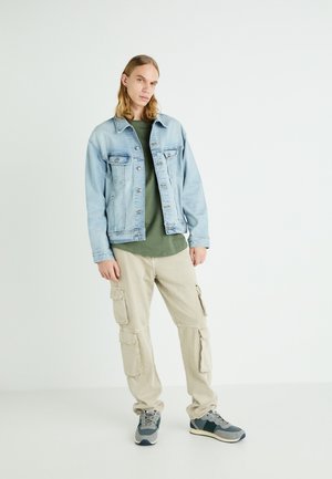 Hellblaues Denim-Jackett, olivgrünes T-Shirt, beige Cargo-Hosen mit Seitentaschen, graue Sneakers mit blauen Akzenten.