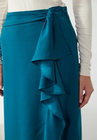 Gonna turchese con dettaglio di ruffle laterale e accentuazione con fiocco. Realizzata in tessuto morbido, presenta una vita aderente e cuciture sottili.