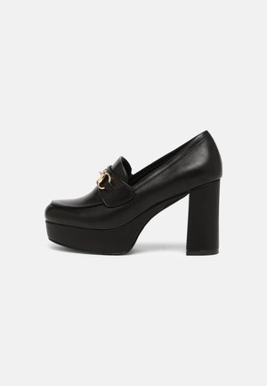 Des mocassins à talons hauts en cuir noir présentent un bout arrondi, une large semelle et une boucle dorée sur le dessus.