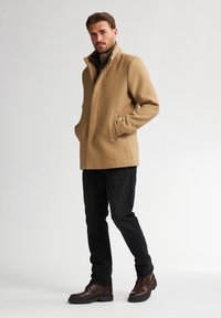 Cappotto di lana beige con colletto alto, due tasche laterali e design minimalista, abbinato a jeans neri e stivaletti stringati marroni. Texture liscia.