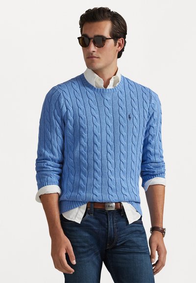 Pull en tricot bleu clair avec un col rond, porté par-dessus une chemise blanche à col, associé à un jean foncé et des lunettes de soleil.