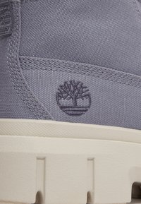 Zapatillas de lona gris con superficie texturizada, que presentan una suela de goma blanca y un logo bordado de un árbol en el lateral.