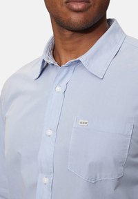 Camicia button-up blu chiaro realizzata in tessuto con una texture liscia. Presenta una tasca sul petto, bottoni bianchi e un'etichetta con il marchio.